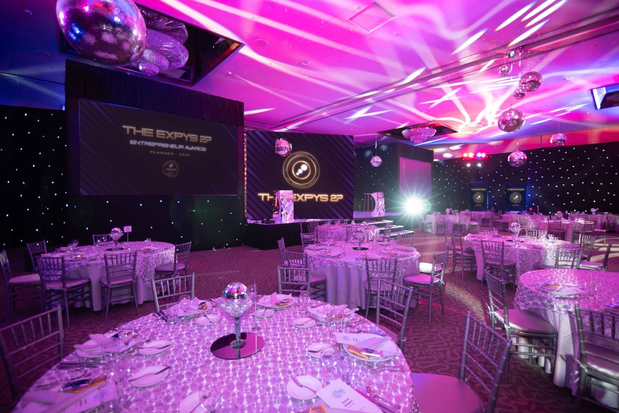 A ballroom dressed for The Expys 27 under pink light, with round tables and the stage screen