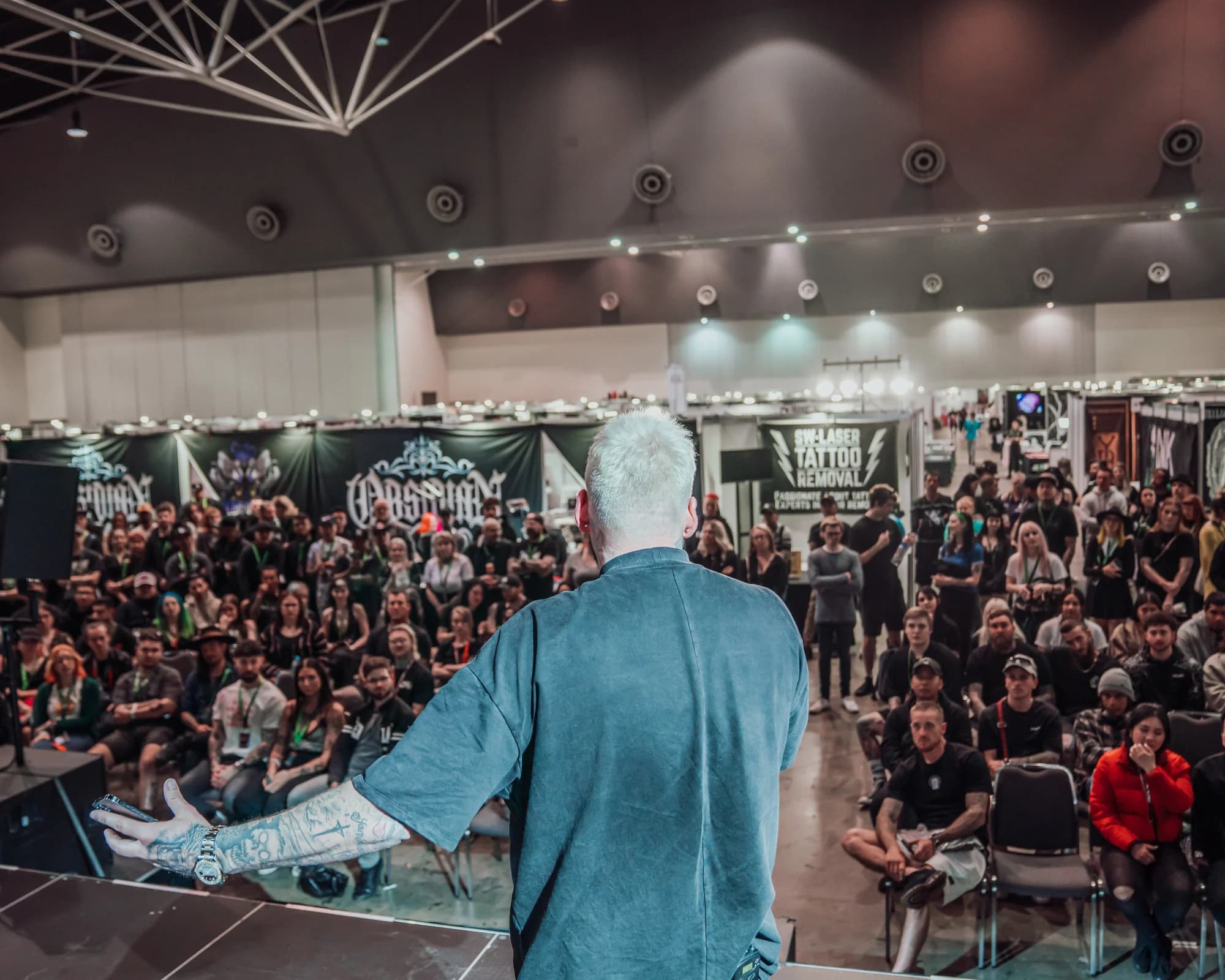 Kevin Mack seen from behind on stage, addressing a seated crowd at an exhibition