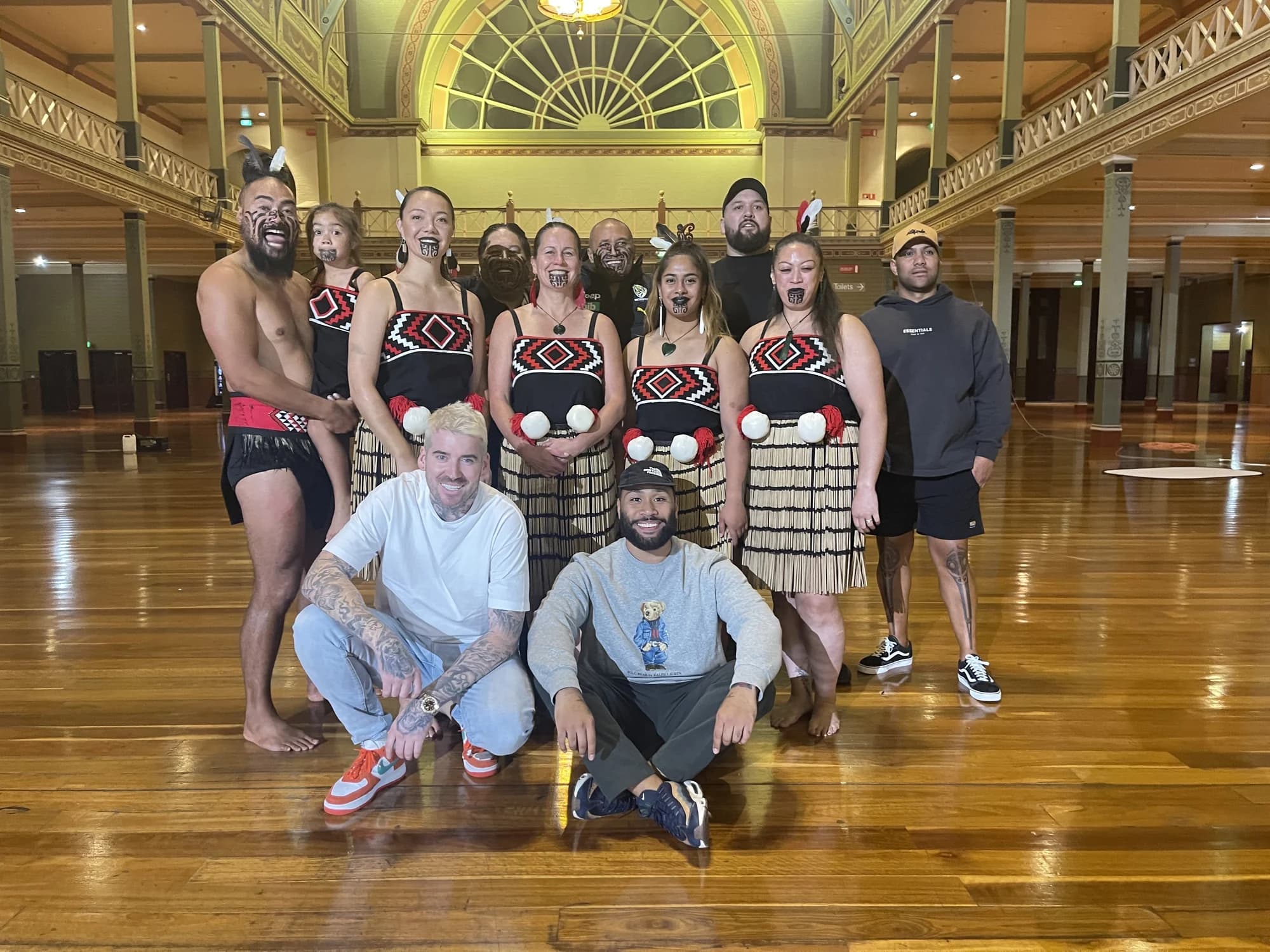 Kevin Mack kneeling with a group of performers in traditional dress inside the Royal Exhibition Building