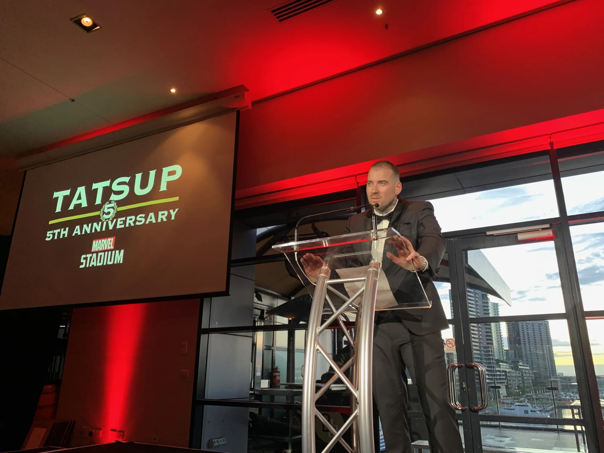 Kevin Mack speaking at a lectern in front of a TatsUp fifth anniversary screen